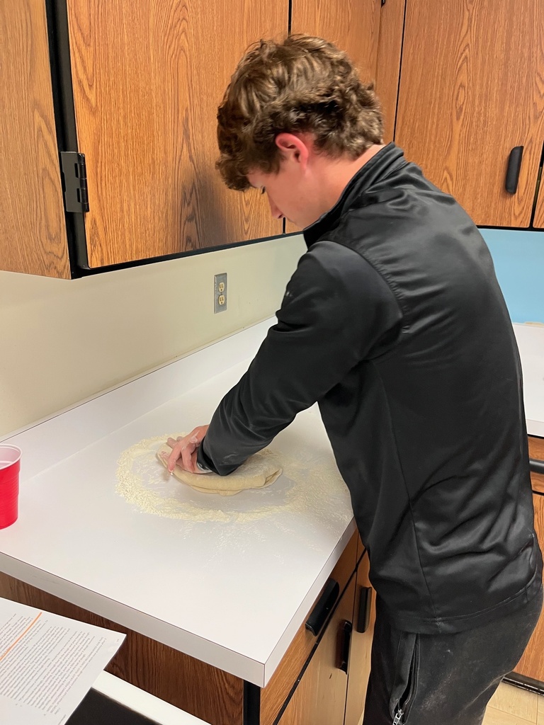 A student is at the counter mixing dough. He is pressing it on a flour base.