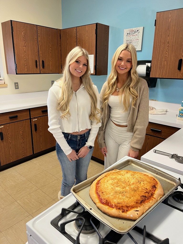 Two students are standing in front of a stove. They  have their pizza on display.