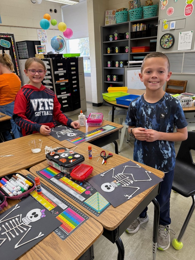 Students are standing at their desks. On the desk are the skeletons they are making.
