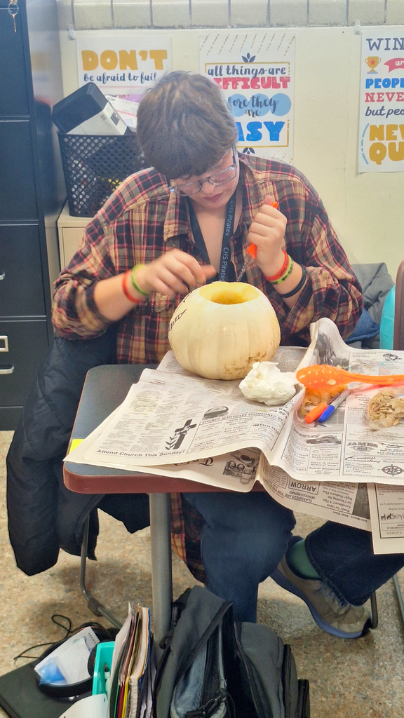 The student is sitting at a table covered in newspaper. She is carving the pumpkin.