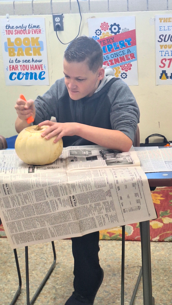 The student is sitting at a table covered in newspaper.  He is carving the pumpkin.