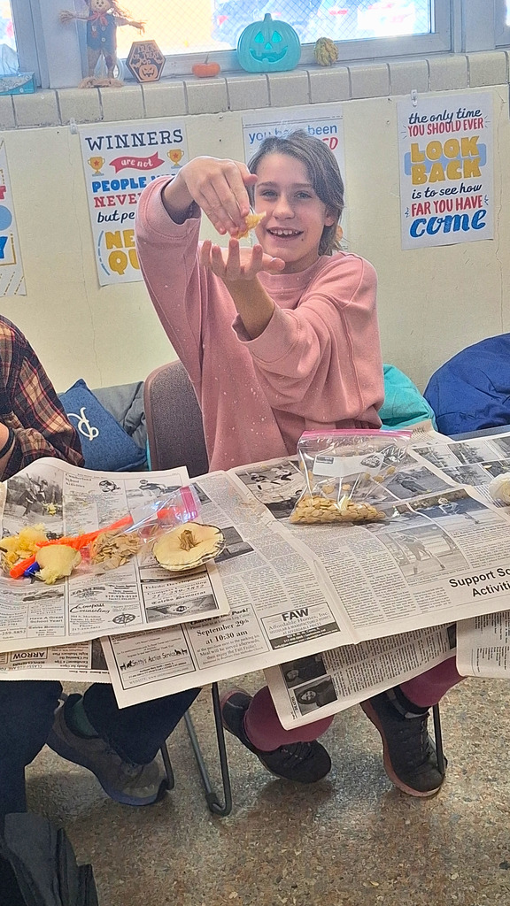 The student is sitting at a table covered in newspaper. She is holding up the pumpkin "guts"!