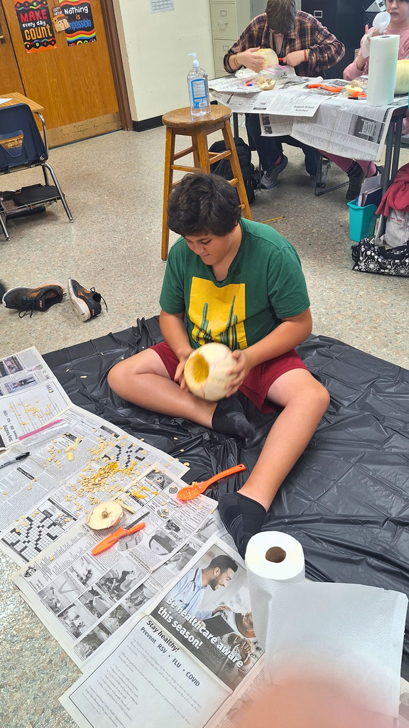 The student is sitting on the floor covered in newspaper. He is emptying a pumpkin of its seeds.