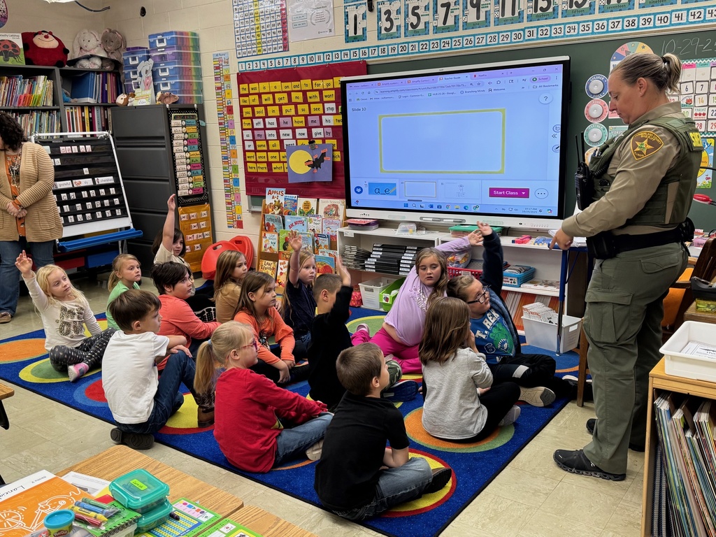 Students are sitting on the class carpet. The are raising their hands. SRO Elizabeth is listening to a student.