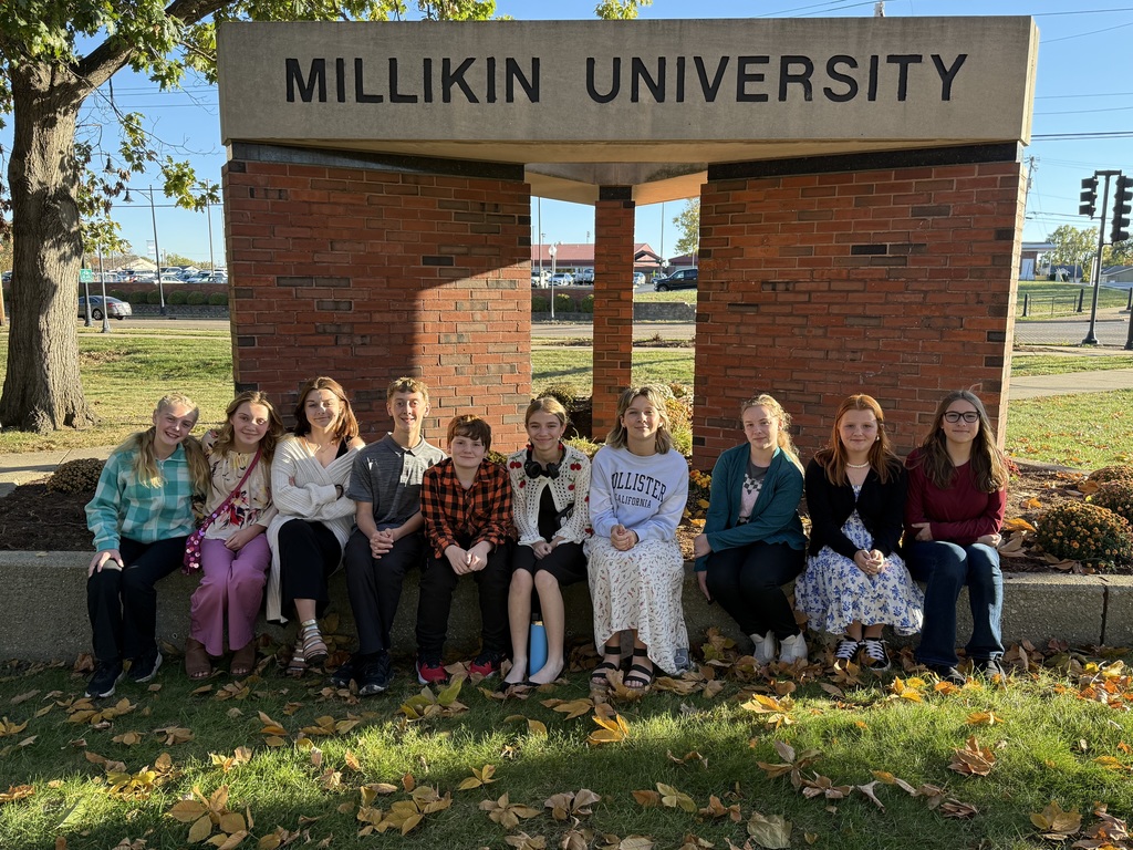 The students are sitting on a concrete wall in front of the Millikin University Sign.
