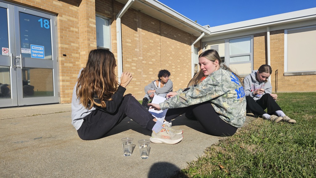 Students are sitting on the ground watching over their cups filled with liquid for their lab.