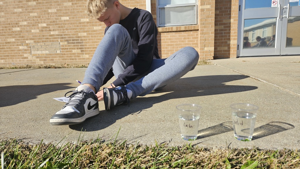 A student is sitting on the ground writing data from his photosynthesis lab. 