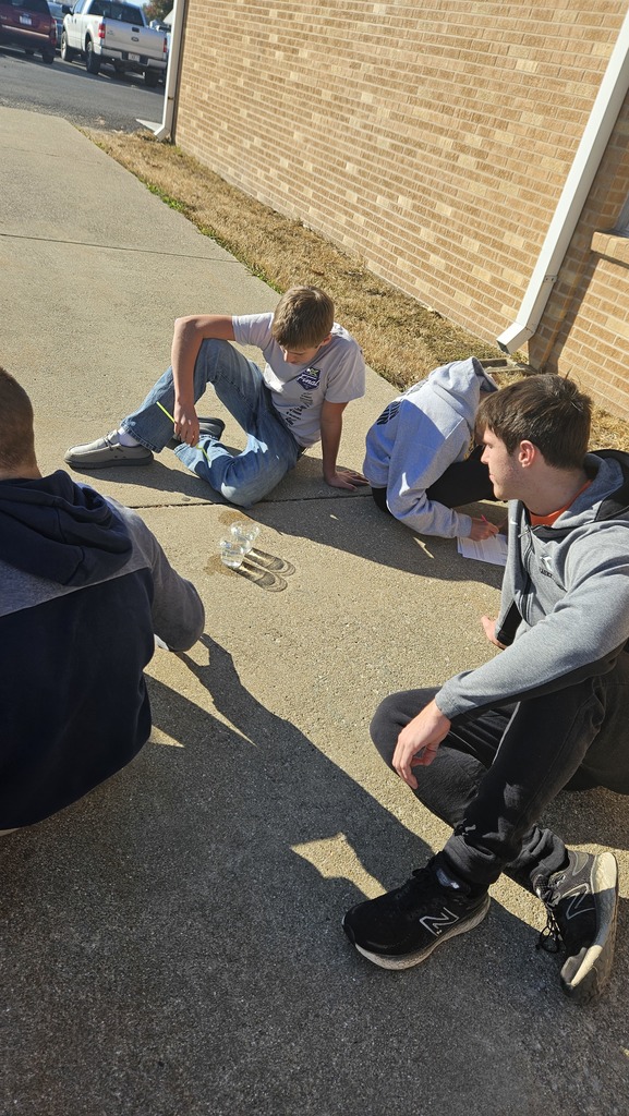 A group of students are sitting on the ground. They are watching the two cups of liquid they have for their lab.