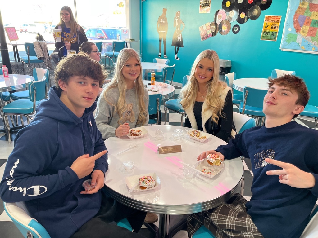 The students are sitting around a table. They have their own decorated donut in front of them. 