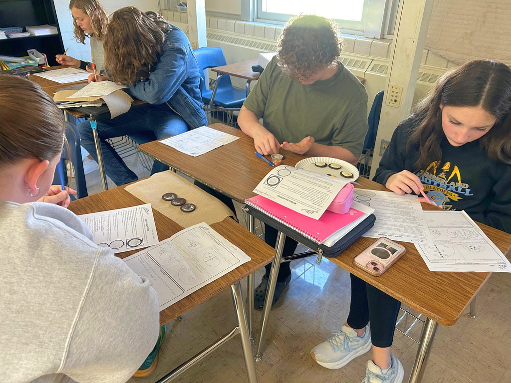Students are sitting at their desks with the cookies and decorating supplies near. The students are using the decorations to create a stage of mitosis. They are also referring to their papers to do the project. 