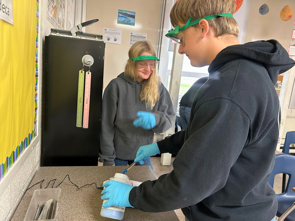 Student are wearing safety goggles and gloves. They are scooping something out of a container.