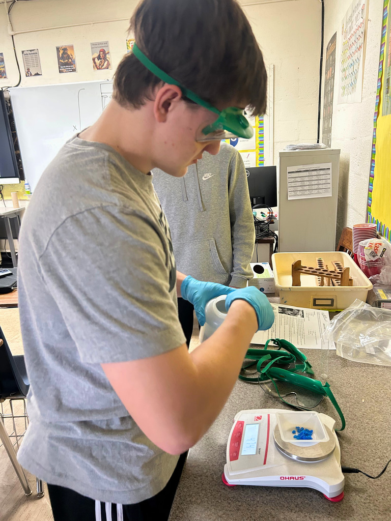 A student is wearing safety goggles and gloves. He is scooping something out of a container.