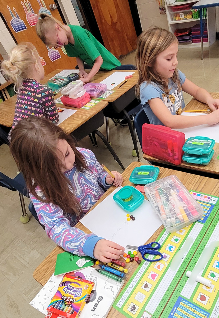Students are sitting at their desks. The image shows a paper with three boxes. There are a number of Skittles in the first box and a different numnber in the second box. The students are to add them together. 