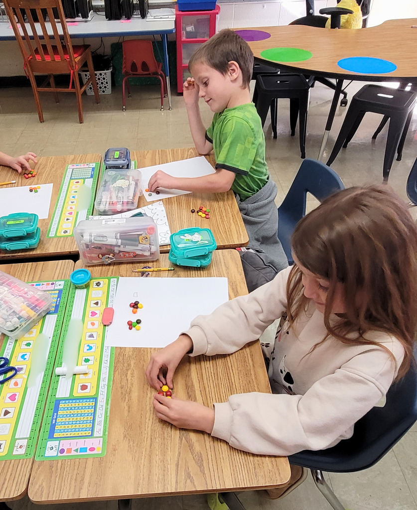 Students are sitting at their desks. The image shows a paper with three boxes. There are a number of Skittles in the first box and a different numnber in the second box. The students are to add them together. 