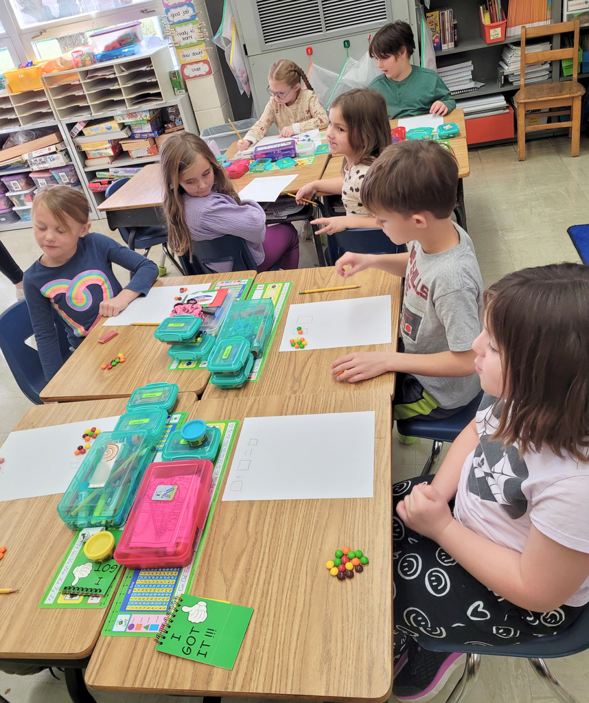 Students are sitting at their desks. The image shows a paper with three boxes. There are a number of Skittles in the first box and a different numnber in the second box. The students are to add them together. 
