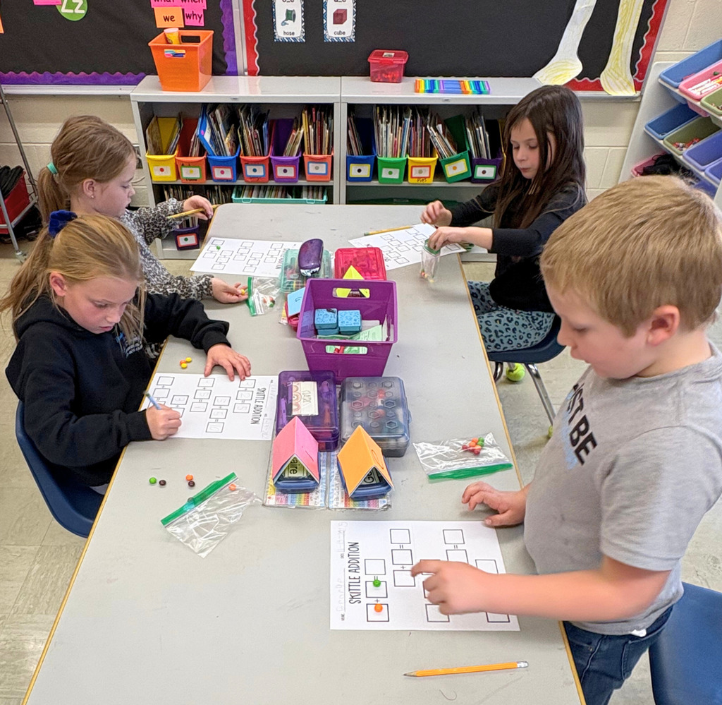 Students are sitting at their desks. The image shows a paper with three boxes. There are a number of Skittles in the first box and a different numnber in the second box. The students are to add them together. 