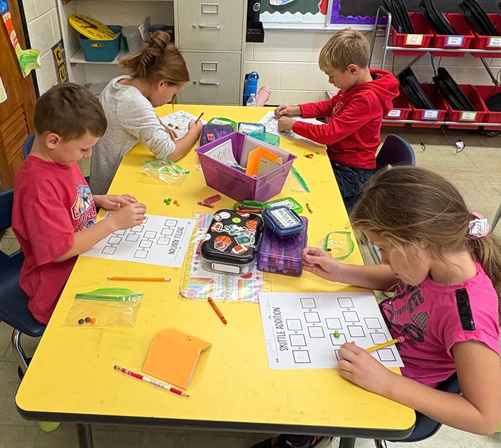 Students are sitting at their desks. The image shows a paper with three boxes. There are a number of Skittles in the first box and a different numnber in the second box. The students are to add them together. 