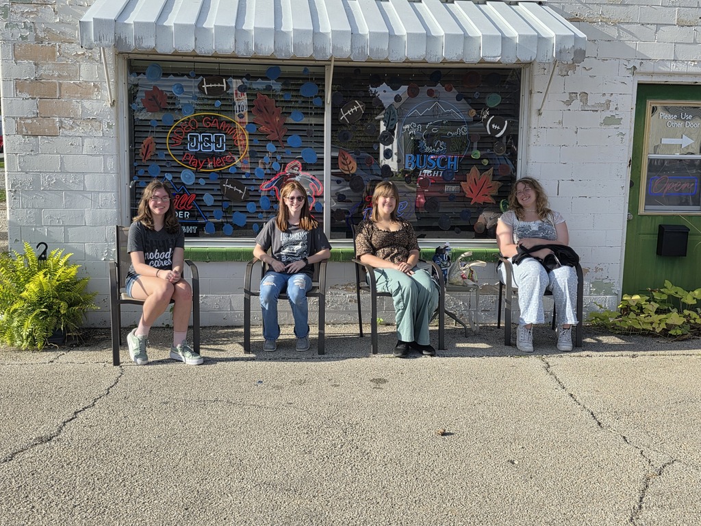 Four students are standing in front of a window that they just painted in a fall theme.