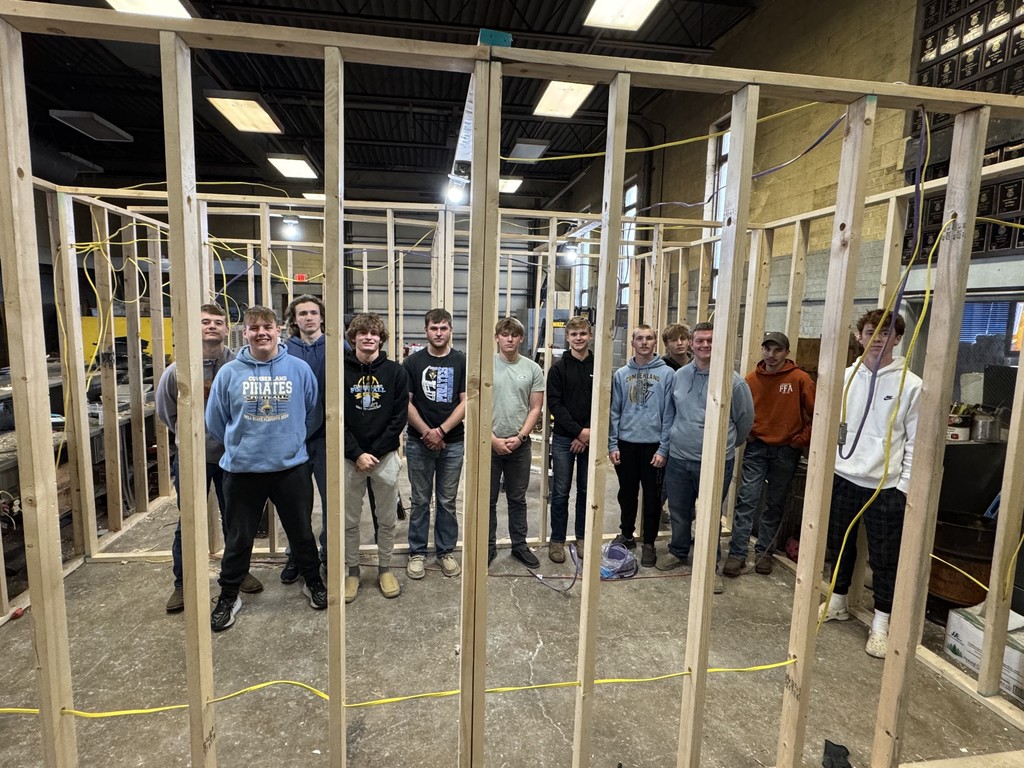 The students are stanidng inside of a makeshift room of studs. They have wired the room for electricity. 