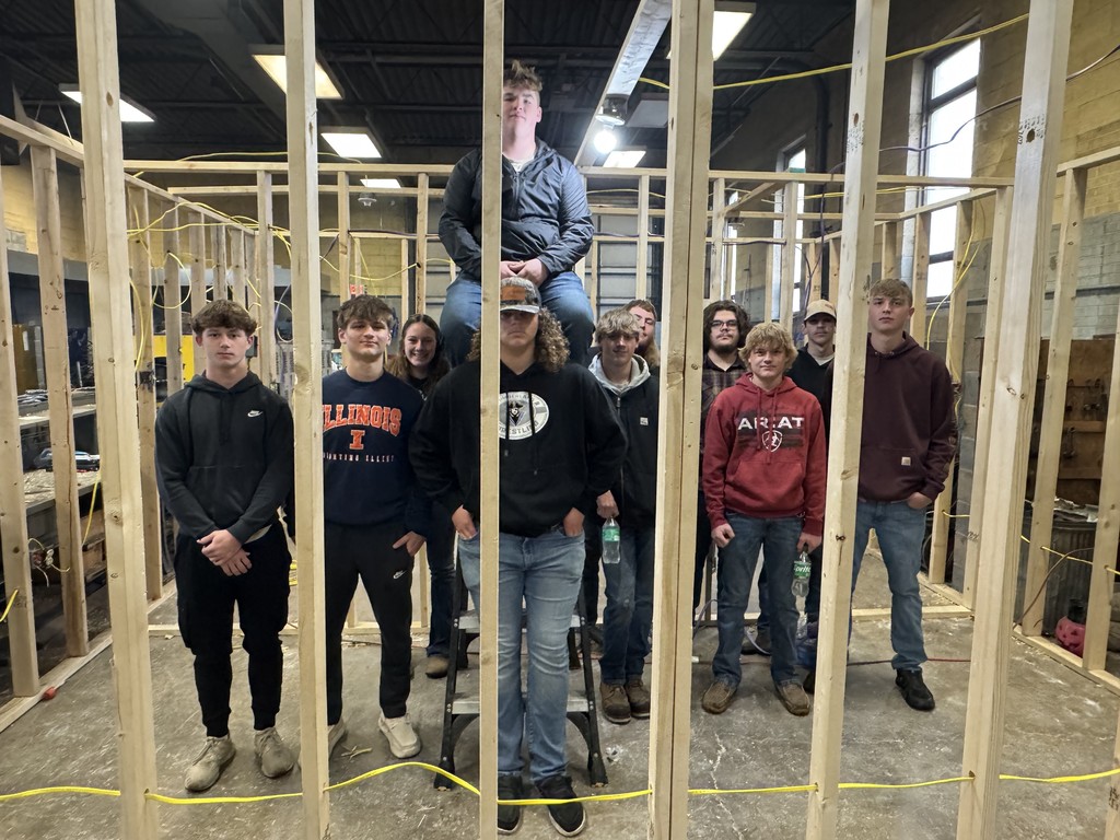 The students are stanidng inside of a makeshift room of studs. They have wired the room for electricity. 