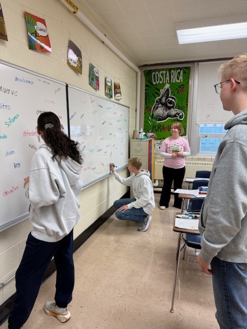 Students are standing at the board conjugating verbs. 