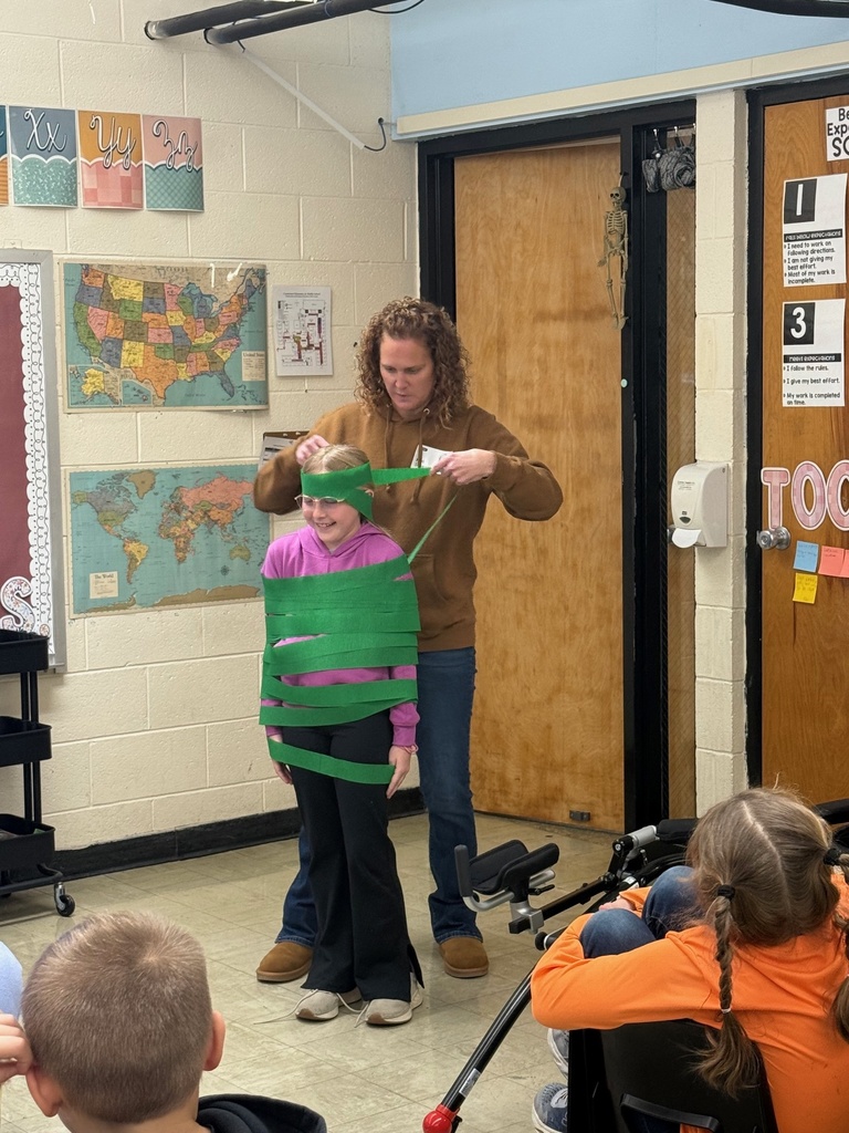 The teacher is wrapping the student with green paper to resemble a tree.