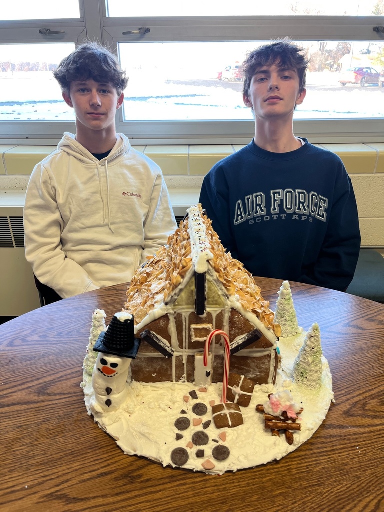 Two studetns are sitting with their completed gingerbread house. It is snow coverd with fortnite accents.