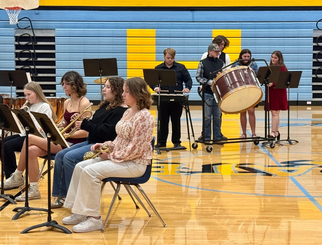 The image shows the brass and percussion sections of the band playing from music on a music stand.