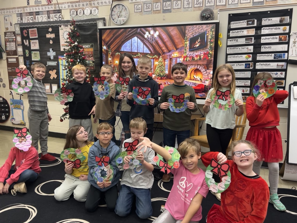 The students are sitting and standing on the carpet. They are holding up their wreaths.
