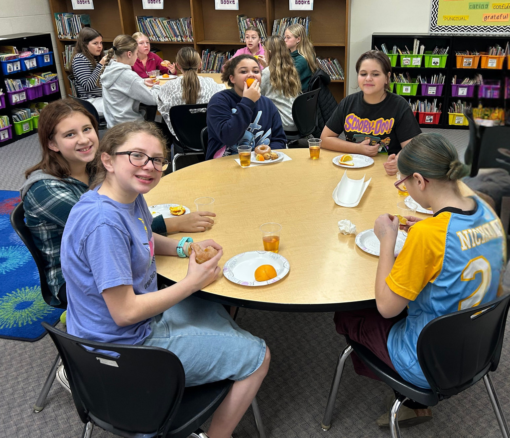 Students are sitting around round tables eating oranges and donuts and drinking apple juice. 