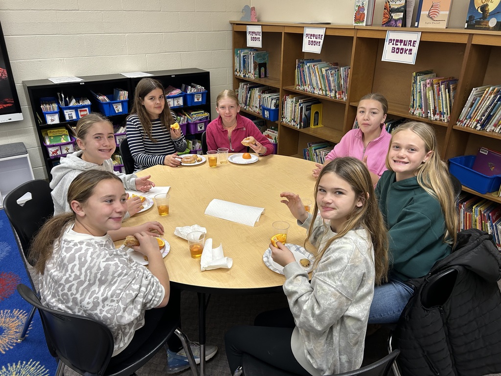 Students are sitting around round tables eating oranges and donuts and drinking apple juice. 