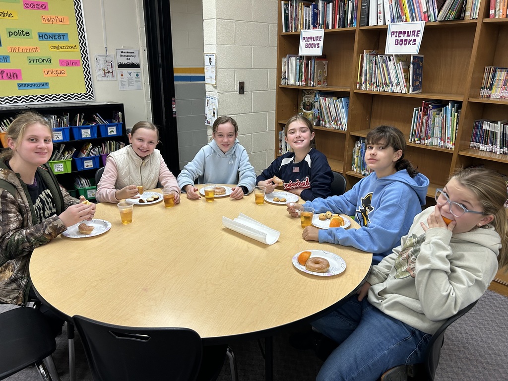 Students are sitting around round tables eating oranges and donuts and drinking apple juice. 