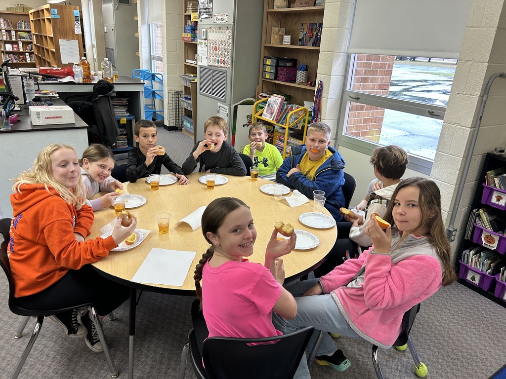 Students are sitting around round tables eating oranges and donuts and drinking apple juice. 
