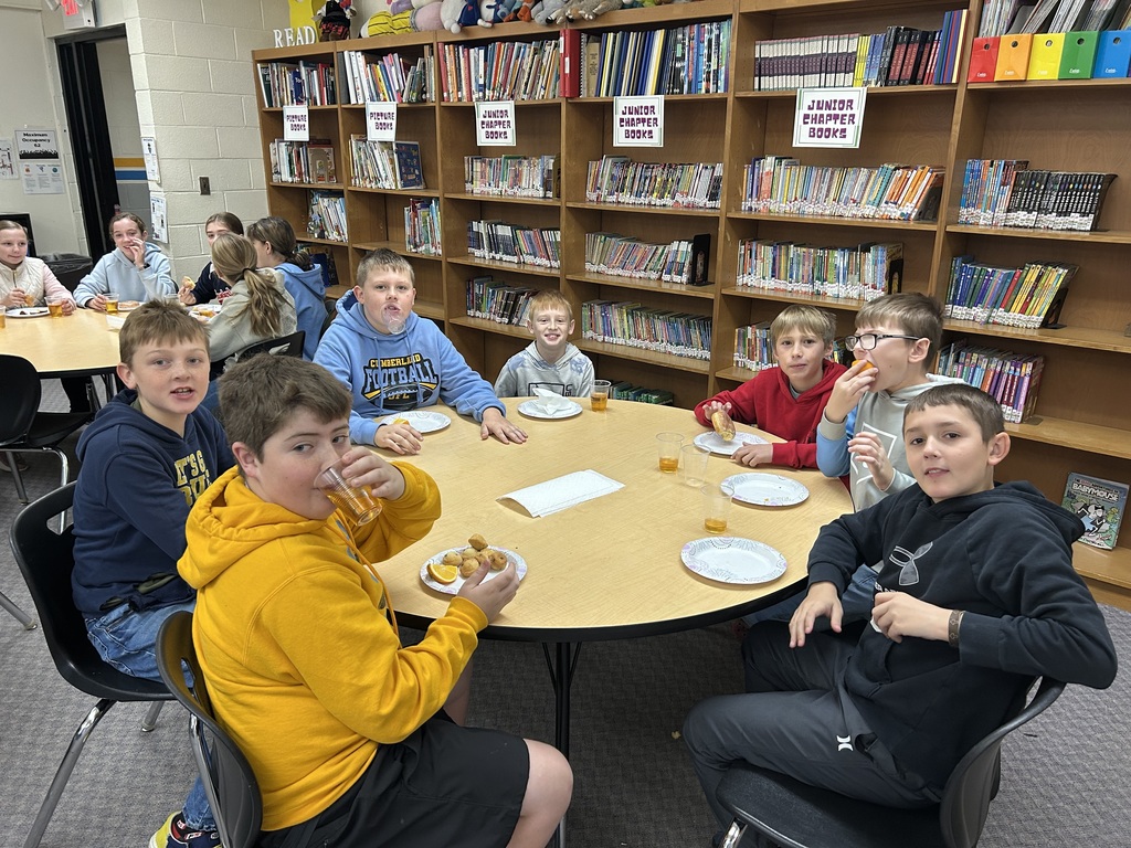 Students are sitting around round tables eating oranges and donuts and drinking apple juice. 