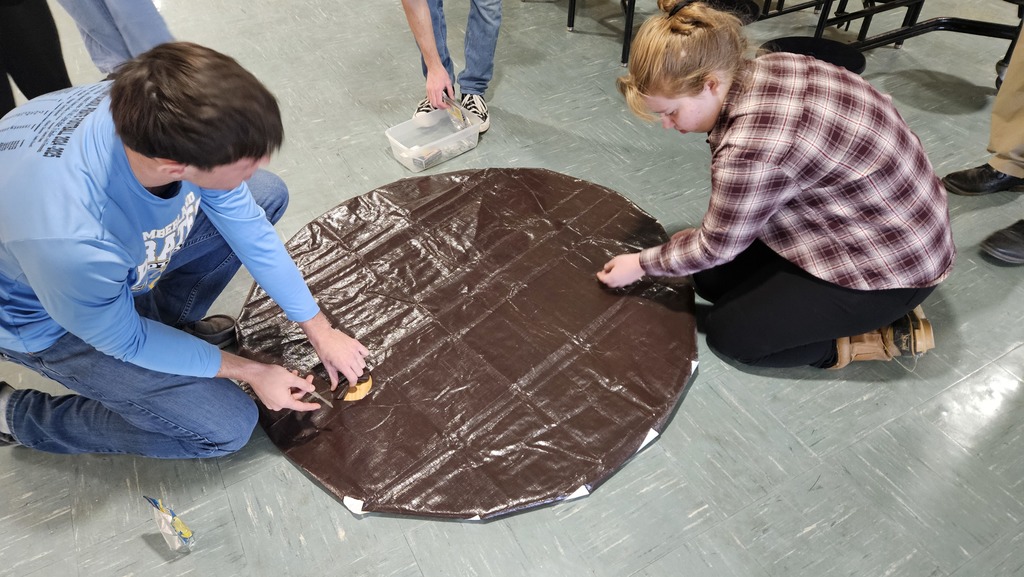The plywood is covered with a tarp. Students are cutting a hole in the tarp.