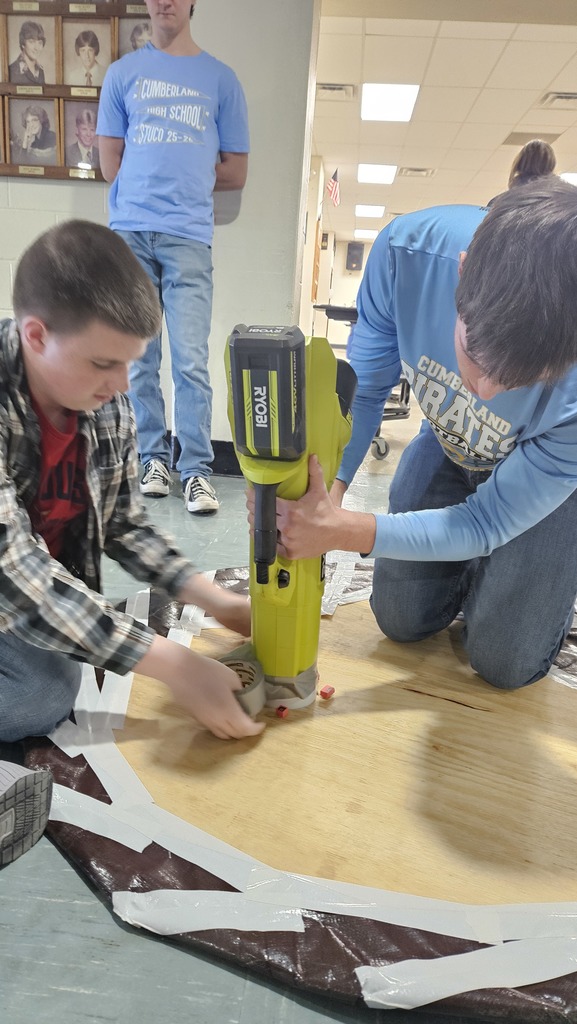 Two students are taping a leaf blower to the hole that has been cut into the plywood.