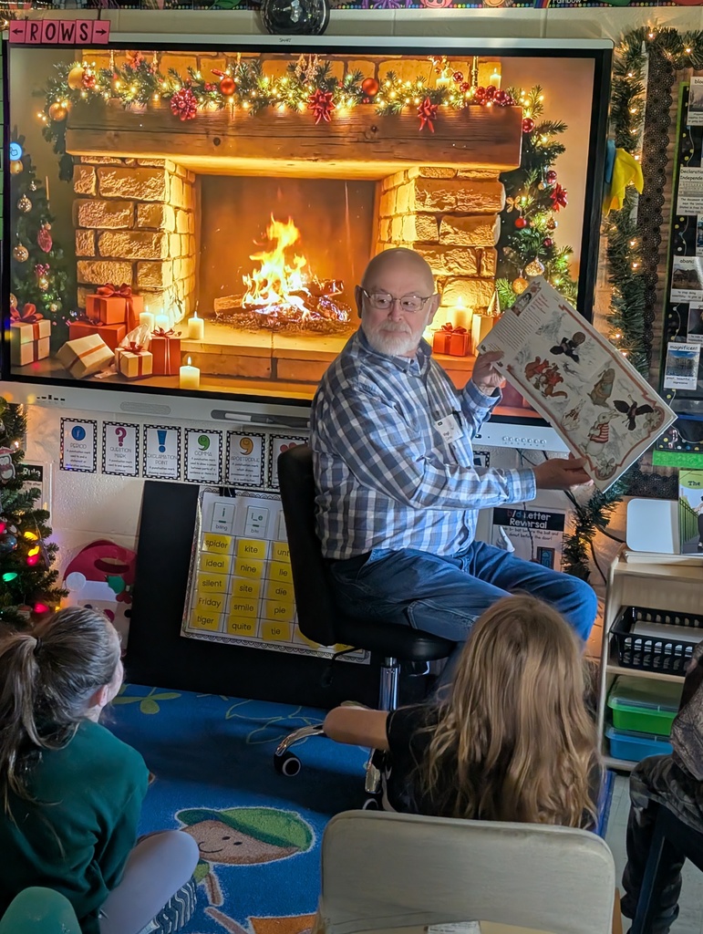 Mr. Virgil is holding up a book to show the pictures to the students. He is sitting in front of a smartboard with a fireplace scene.
