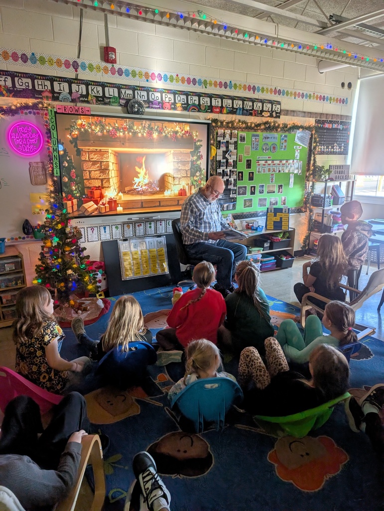 Mr. Virgil is reading a book to the students. He is sitting in front of a smartboard with a fireplace scene. The students are sitting in chairs and on the carpet.