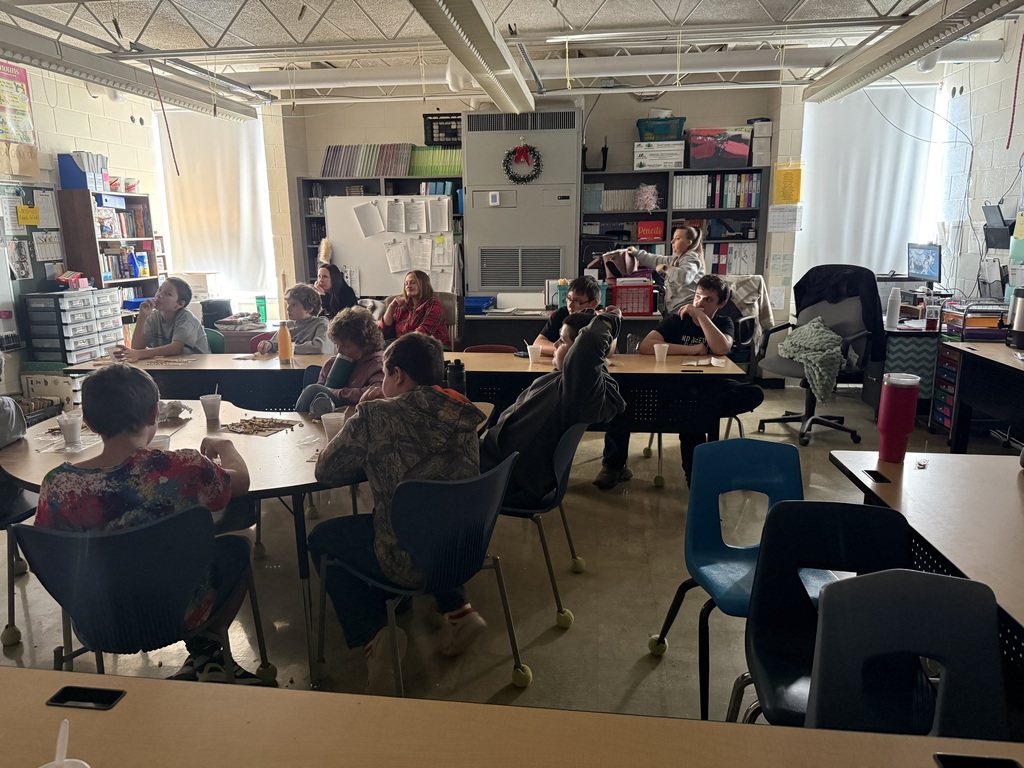 The students are sitting in a darkened room, eating a snack and drinking hot cocoa, They are watching a movie.