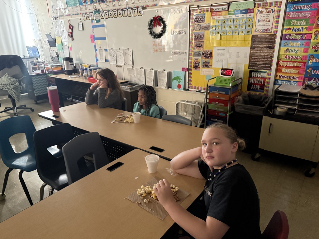 The students are sitting in a darkened room, eating a snack and drinking hot cocoa, They are watching a movie.