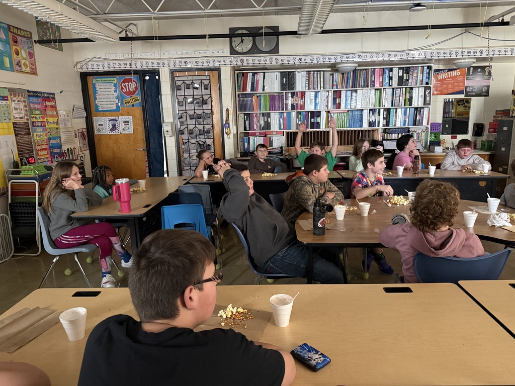 The students are sitting in a darkened room, eating a snack and drinking hot cocoa, They are watching a movie.
