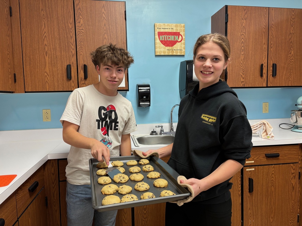 Two students are standing at the counter holding a pan of baked chocolate chip cookies. One student is using a spatula to serve a cookie. 