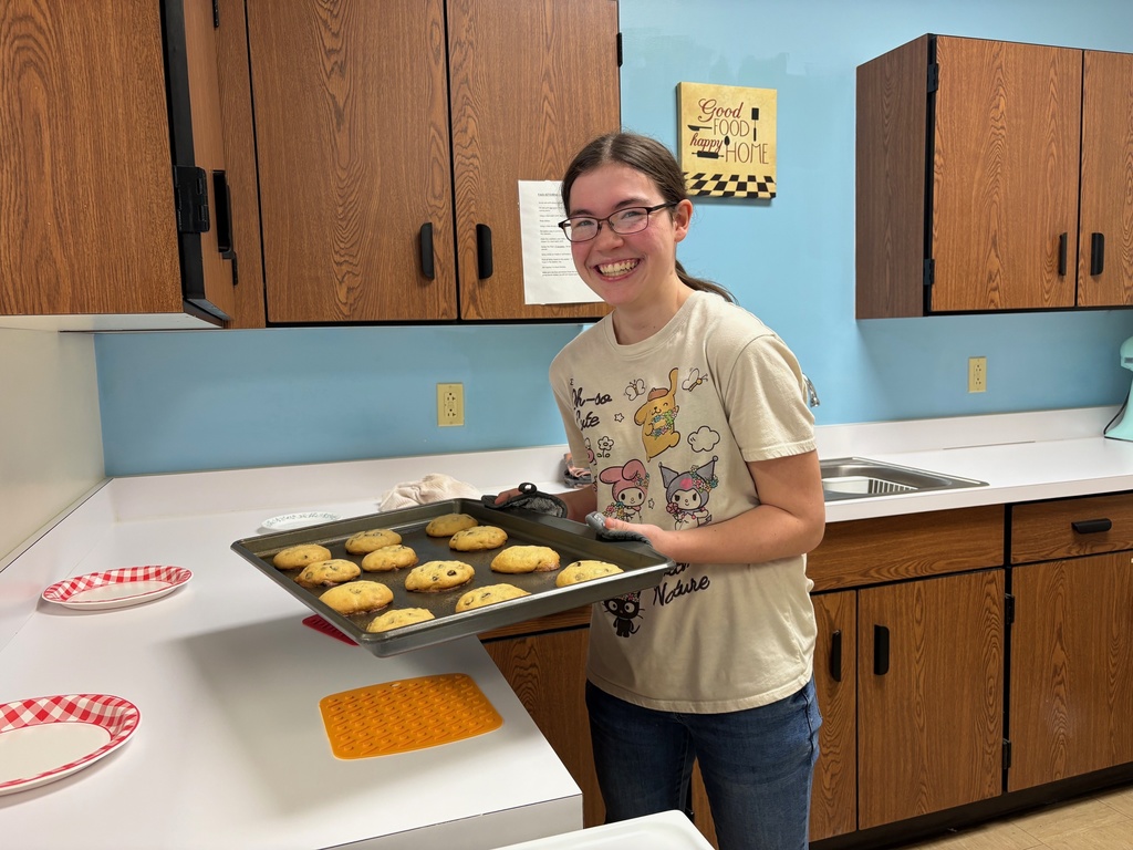 A student is standing at the counter holding a pan of  baked chocolate chip cookies. 