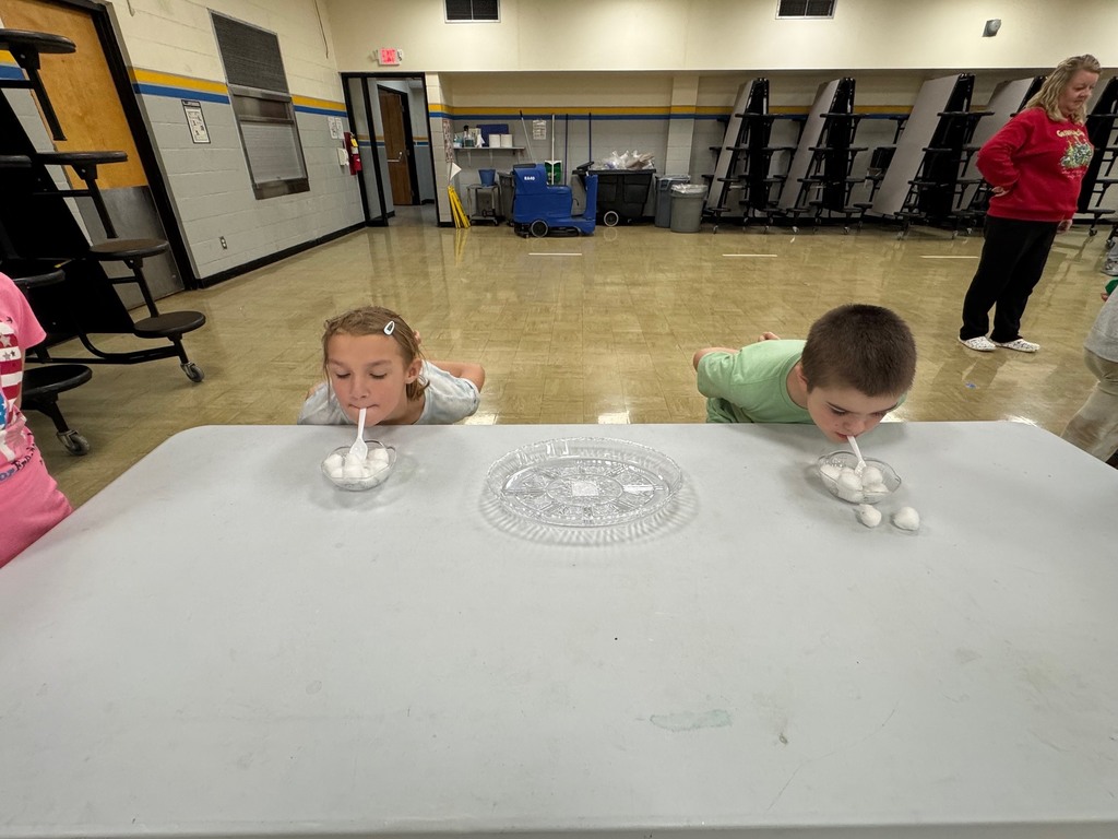 Students have a spoon in their mouths. They have to move the cotton balls from one dish to the other using only the spoon and no hands. 