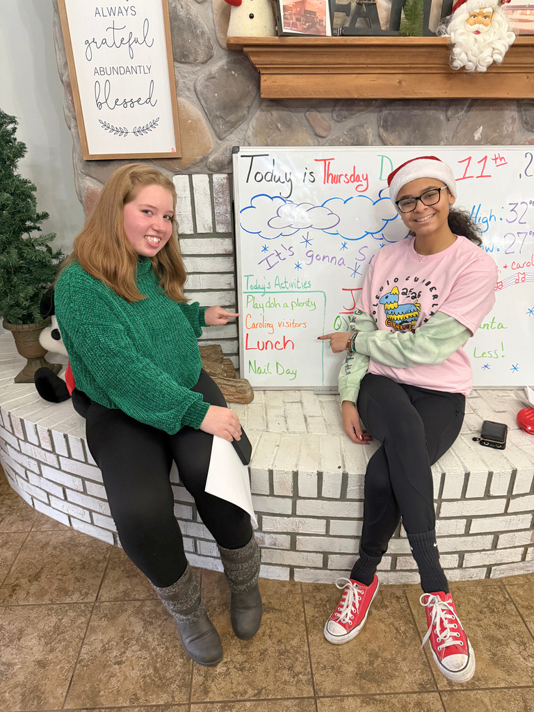Students are sitting at a fireplace. They are pointing to their group's name on the white board. 