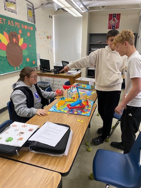 Three students are using the Mouse Trap game setup to identify types of physical science. They are sitting around a table with the game in the middle. .