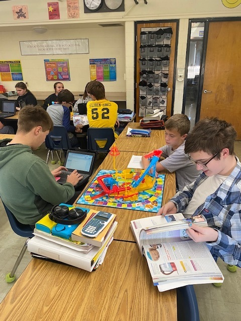 Three students are using the Mouse Trap game setup to identify types of physical science. They are sitting around a table with the game in the middle. .