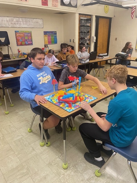 Three students are using the Mouse Trap game setup to identify types of physical science. They are sitting around a table with the game in the middle. .