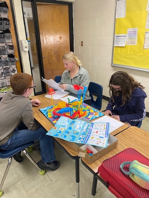 Three students are using the Mouse Trap game setup to identify types of physical science. They are sitting around a table with the game in the middle. .