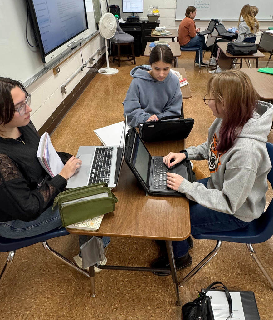 A small group of students are sitting around the desks. They are researching on their Chromebooks. and discussing. 